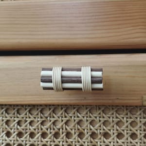 Può includere: Una maniglia per cassetto orizzontale composta da aste in legno alternate in marrone scuro e beige chiaro, tenute insieme da due fasce in rattan intrecciato di colore chiaro. La maniglia è montata su un frontale in legno chiaro con pannello in paglia.