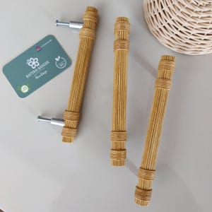May include: Three natural rattan cabinet pulls with silver fittings. The pulls have a cylindrical, ribbed design and are arranged on a light surface. A tag with the text "RATTAN SANDIK Home Design" is also present.
