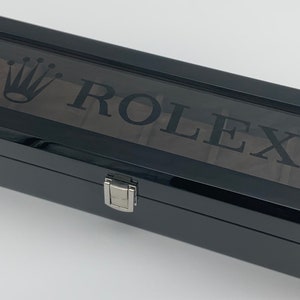 May include: A black rectangular watch box with a clear glass top. The box has a silver latch and the words "ROLEX" and a crown logo are printed on the top.
