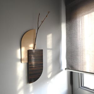 Könnte beinhalten: Eine moderne Wandvase aus Holz mit natürlicher Holzoberfläche und einer dunklen, gestreiften Holzvase. Ein schlanker Zweig mit kleinen Knospen befindet sich in der Vase. Die Vase ist an einer hellgrauen Wand befestigt.