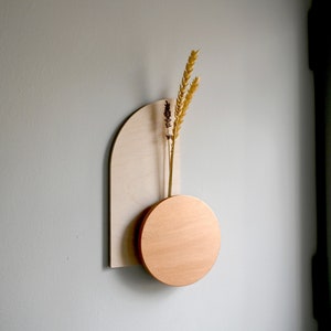 May include: A modern wall-mounted vase with a light wood arch and a copper-colored circular base. The vase holds dried wheat stalks. The design is minimalist and geometric, suitable for home decor.
