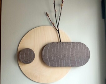 May include: A modern wall art piece featuring a light wood circular base with a smaller circular accent and an oblong accent in a gray wood grain pattern. The art holds a few slender branches with small, light-colored buds.