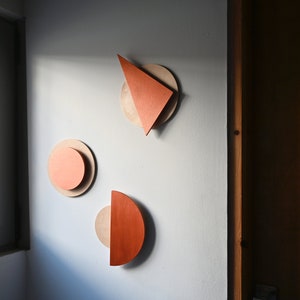 Op de afbeelding: Drie wanddecoratie-objecten met gelaagde geometrische vormen. Elk stuk bestaat uit een lichtbeige cirkelvormige basis met daarop een kleiner, feloranje element in de vorm van een driehoek, een cirkel of een halve cirkel.