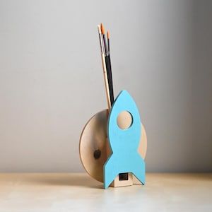 Könnte beinhalten: Ein hölzerner Raketen-Pinselhalter mit einer blauen Rakete und einem braunen Holzsockel. Der Halter steht auf einem Holztisch.