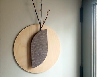 May include: A wooden wall art piece featuring a light brown circular base with a gray, leaf-shaped accent. Dark branches with small, light-colored buds extend from a gray vase-like structure, creating a natural, minimalist aesthetic.