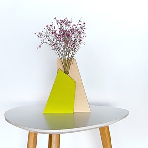 Könnte beinhalten: Eine Holzvase mit geometrischem Design in Beige- und Limettentönen, die einen Strauß rosa Blumen hält. Die Vase steht auf einem weißen Tisch mit Holzbeinen.