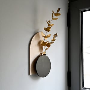 May include: A black circular vase attached to a light wood arch-shaped wall decor. A sprig of dried golden leaves is in the vase.