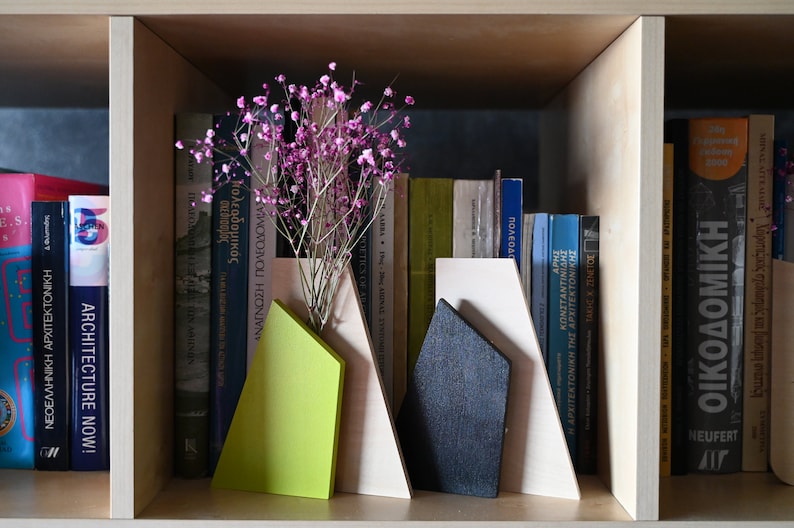 K&ouml;nnte beinhalten: Ein Satz aus drei Holzbuchst&uuml;tzen in Form von Bergen. Die Buchst&uuml;tzen sind in Beige-, Gr&uuml;n- und Schwarzt&ouml;nen lackiert. Die Buchst&uuml;tzen stehen in einem B&uuml;cherregal mit einer Auswahl an B&uuml;chern und einer Vase mit rosa Blumen.