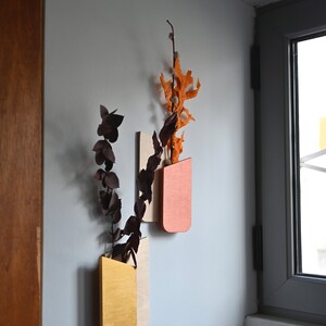 K&ouml;nnte beinhalten: Zwei wandmontierte Holzvase mit abgerundetem Boden, eine in Senfgelb und die andere in Terrakotta-Orange lackiert. Jede Vase enth&auml;lt getrocknete botanische St&auml;ngel, darunter dunkelviolette Eukalyptusbl&auml;tter und orangefarbene Eichenbl&auml;tter.