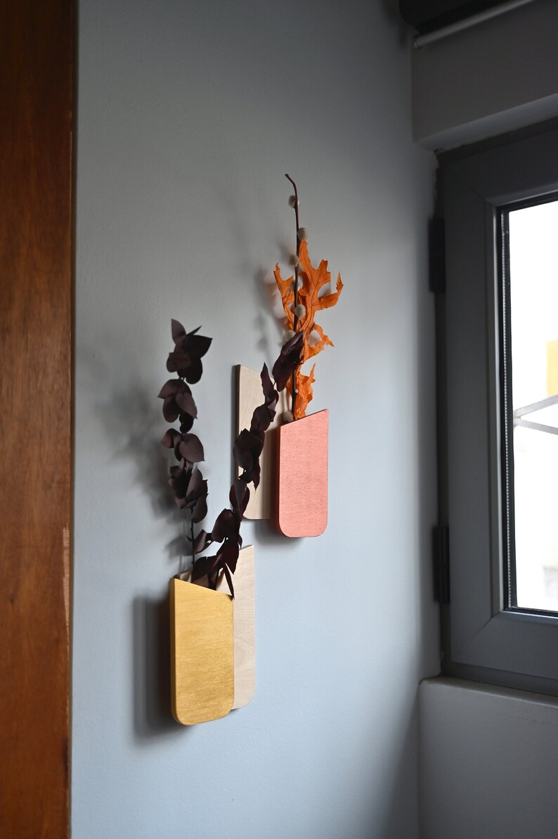 K&ouml;nnte beinhalten: Zwei wandmontierte Holzvase mit abgerundetem Boden, eine in Senfgelb und die andere in Terrakotta-Orange lackiert. Jede Vase enth&auml;lt getrocknete botanische St&auml;ngel, darunter dunkelviolette Eukalyptusbl&auml;tter und orangefarbene Eichenbl&auml;tter.