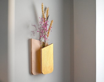 May include: A wall-mounted wooden vase featuring a natural light-colored plywood back panel and a front section finished in metallic gold paint. The vase holds a small arrangement of dried pink flowers and golden wheat stalks.