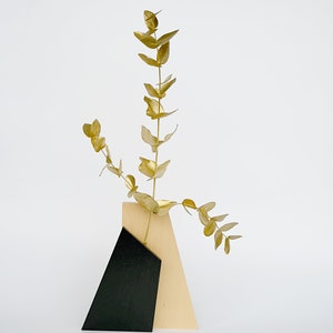 K&ouml;nnte beinhalten: Eine Holzvase mit geometrischem Design in Schwarz und Naturholz enth&auml;lt einen Zweig goldenen Eukalyptus.