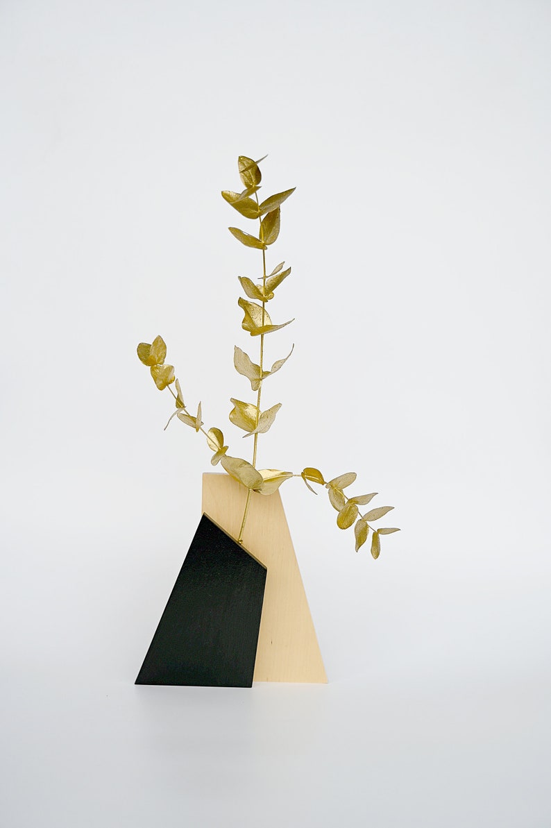 K&ouml;nnte beinhalten: Eine Holzvase mit geometrischem Design in Schwarz und Naturholz enth&auml;lt einen Zweig goldenen Eukalyptus.