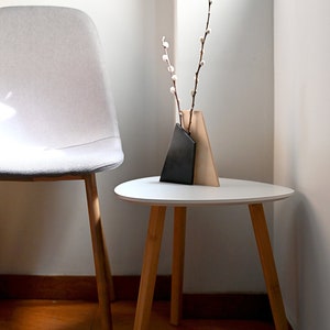 K&ouml;nnte beinhalten: Ein wei&szlig;er Beistelltisch mit einer abgerundeten, ovalen Tischplatte und drei Holzbeinen. Eine kleine Vase mit geometrischem Design in Schwarz und Beige h&auml;lt einen Zweig wei&szlig;er Weidenk&auml;tzchen.
