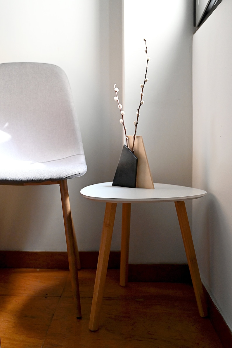 K&ouml;nnte beinhalten: Ein wei&szlig;er Beistelltisch mit einer abgerundeten, ovalen Tischplatte und drei Holzbeinen. Eine kleine Vase mit geometrischem Design in Schwarz und Beige h&auml;lt einen Zweig wei&szlig;er Weidenk&auml;tzchen.