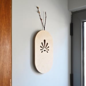 May include: A light wood wall-mounted vase with a cutout design in the shape of a fan. The vase is oval-shaped and has a natural wood finish.