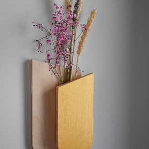 K&ouml;nnte beinhalten: Eine wandmontierte Holzvase mit einer R&uuml;ckwand aus hellem Natur-Sperrholz und einer Vordertasche in Metallic-Gold. Die Vase enth&auml;lt ein Trockenblumenarrangement aus rosa Schleierkraut, Weizen&auml;hren und Lavendel.