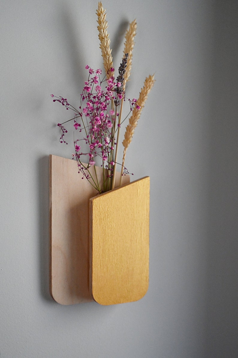 K&ouml;nnte beinhalten: Eine wandmontierte Holzvase mit einer R&uuml;ckwand aus hellem Natur-Sperrholz und einer Vordertasche in Metallic-Gold. Die Vase enth&auml;lt ein Trockenblumenarrangement aus rosa Schleierkraut, Weizen&auml;hren und Lavendel.
