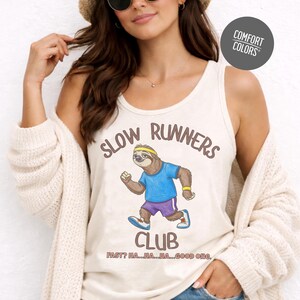 May include: Cream-colored tank top with the words "SLOW RUNNERS CLUB" and a cartoon sloth running. The sloth wears a blue shirt, purple shorts, and a yellow headband. The text "FAST? HA...HA...HA...GOOD ONE." is below the sloth.