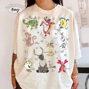 Puede incluir: Camiseta marfil Comfort Colors con personajes de dibujos animados coloridos, incluyendo a Pascal, Mushu, Flounder, Abu, Lumiere, Olaf, Chip, Sven y Sebastián. La camiseta tiene el texto "Comfort Color Ivory" en la parte superior.