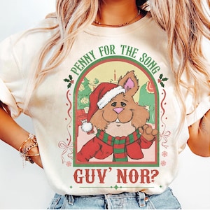 Può includere: T-shirt color crema con un coniglio dei cartoni animati con cappello e sciarpa di Babbo Natale, con la scritta "PENNY FOR THE SONG GUY NOR?" in un design festivo. Il coniglio è ambientato su uno sfondo a tema natalizio.