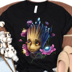 May include: Black t-shirt featuring a colorful illustration of a Groot-like character surrounded by vibrant flowers. The artwork showcases a detailed design with shades of brown, blue, and pink, creating a whimsical and eye-catching graphic. The shirt is a classic crew neck.