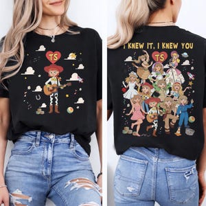 May include: A black t-shirt featuring a front graphic of a cowgirl character playing a guitar with a heart containing the letters TS. The back graphic displays multiple female characters in various outfits, the text I KNEW IT, I KNEW YOU, and a heart with TS.