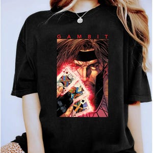 Può includere: T-shirt nera con un'illustrazione a fumetti di Gambit, che tiene in mano delle carte da gioco con un effetto energia rosso. La parola "GAMBIT" è stampata sopra l'illustrazione. La maglietta è in materiale morbido.