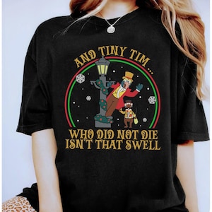 Puede incluir: Camiseta negra con una ilustración de dibujos animados. El diseño incluye el texto "AND TINY TIM... WHO DID NOT DIE ISN'T THAT SWELL" y un personaje con abrigo rojo y sombrero de copa, y una farola.