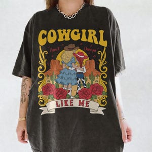 K&ouml;nnte beinhalten: Ein anthrazitfarbenes Kurzarm-T-Shirt mit einem Grafikdruck von zwei Figuren in einer W&uuml;stenlandschaft mit Kakteen, roten Rosen und dem Text COWGIRL I knew it I knew you LIKE ME in gelber, roter und wei&szlig;er Schrift auf der Vorderseite.