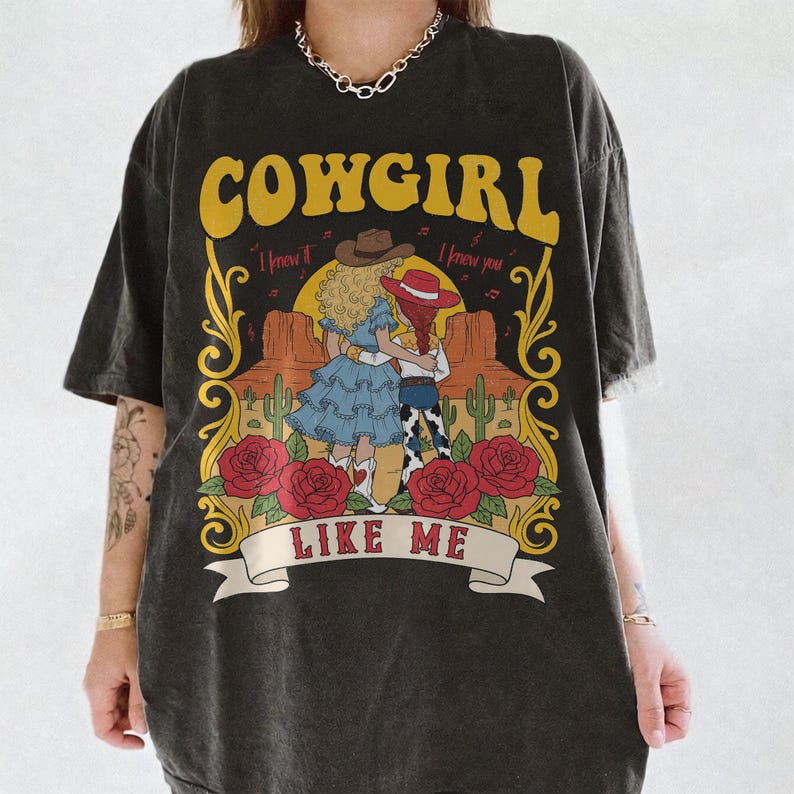 K&ouml;nnte beinhalten: Ein anthrazitfarbenes Kurzarm-T-Shirt mit einem Grafikdruck von zwei Figuren in einer W&uuml;stenlandschaft mit Kakteen, roten Rosen und dem Text COWGIRL I knew it I knew you LIKE ME in gelber, roter und wei&szlig;er Schrift auf der Vorderseite.