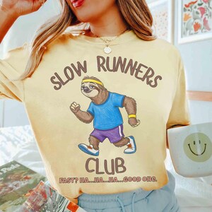 May include: Yellow t-shirt with the text "SLOW RUNNERS CLUB" and a cartoon sloth wearing a headband, blue shirt, and purple shorts. The text "FAST? HA...HA...HA...GOOD one." is below the club name.