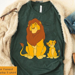 May include: A dark green Bella T-shirt featuring a cartoon illustration of a lion and a cub. The lion has a brown mane and the cub is yellow. The text "Heather Forest Only Bella T-shirt" is in a yellow box.