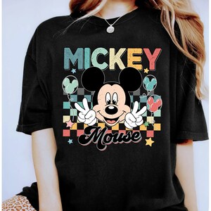 K&ouml;nnte beinhalten: Schwarzes T-Shirt mit einer Retro-Grafik von Mickey Mouse mit dem Text "Mickey Mouse" in Regenbogenfarben.