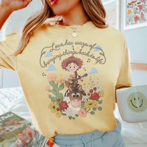 Pu&ograve; includere: Una t-shirt giallo pallido con la grafica di una cowgirl dei cartoni animati che suona la chitarra su un cavallo. Il design include cactus, fiori colorati, note musicali e il testo "Love has ways of bringing things back to life."