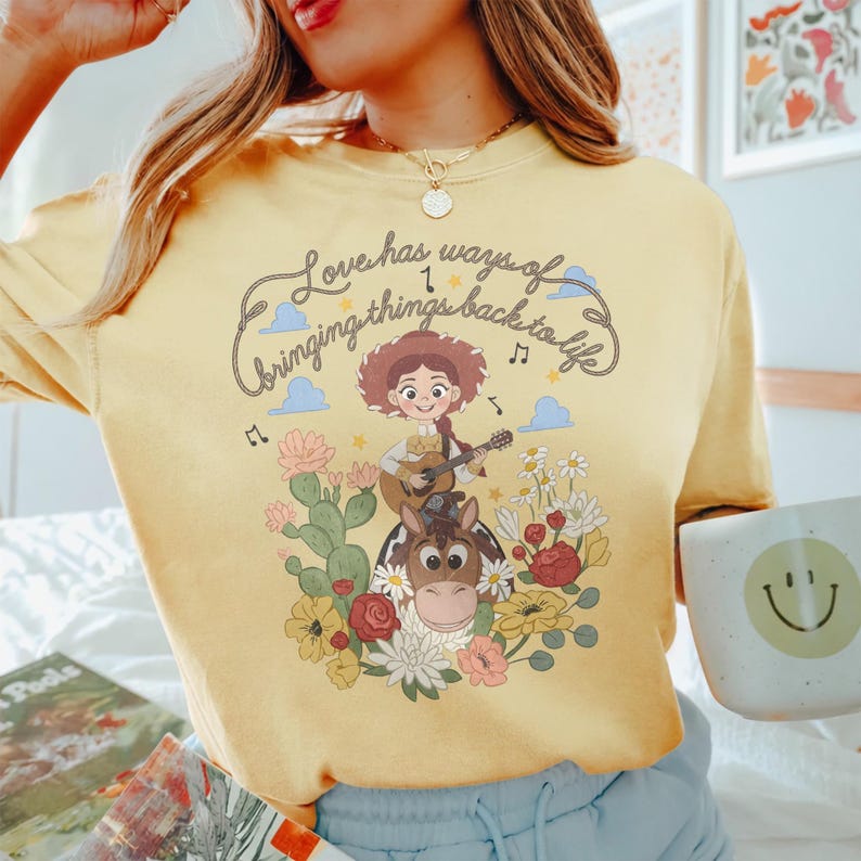 Pu&ograve; includere: Una t-shirt giallo pallido con la grafica di una cowgirl dei cartoni animati che suona la chitarra su un cavallo. Il design include cactus, fiori colorati, note musicali e il testo "Love has ways of bringing things back to life."