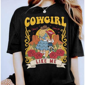 K&ouml;nnte beinhalten: Ein schwarzes T-Shirt mit einem Grafikdruck von zwei Figuren in Westernkleidung in einer W&uuml;stenlandschaft mit Kakteen und roten Rosen. Der Text lautet COWGIRL LIKE ME mit dem Schriftzug I knew it I knew you &uuml;ber den Figuren.