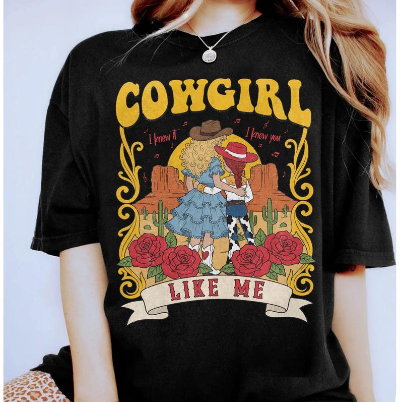 K&ouml;nnte beinhalten: Ein schwarzes T-Shirt mit einem Grafikdruck von zwei Figuren in Westernkleidung in einer W&uuml;stenlandschaft mit Kakteen und roten Rosen. Der Text lautet COWGIRL LIKE ME mit dem Schriftzug I knew it I knew you &uuml;ber den Figuren.