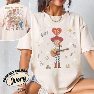 May include: An ivory t-shirt featuring a graphic of a cowgirl character playing an acoustic guitar. Above the character is a red heart with the letters TS. The design includes musical notes, clouds, stars, a rocket, and a ball. The back of the shirt shows a larger collage of various characters with the text I KNEW IT, I KNEW YOU TS.