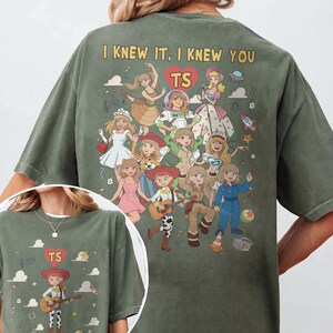 May include: An olive green t-shirt featuring a graphic print on the back and front. The back displays multiple cartoon characters in various outfits with the text "I KNEW IT, I KNEW YOU" and a heart containing "TS". The front shows one character.
