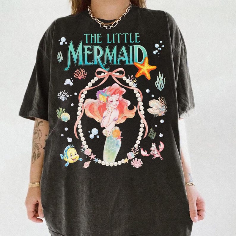 以下が含まれることがあります： 黒いTシャツ。リトルマーメイドのアリエルのカラフルなグラフィックが特徴です。「THE LITTLE MERMAID」の文字はティールと白で、海の生き物と真珠で縁取られたアリエルの肖像画のイラストが描かれています。