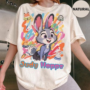 May include: Cream-coloured t-shirt featuring a colourful graphic of Judy Hopps from Zootopia. The design includes Judy, a bunny, with the text "Judy Hopps" in a vibrant font. Additional cartoon characters and colourful paint strokes complete the design.