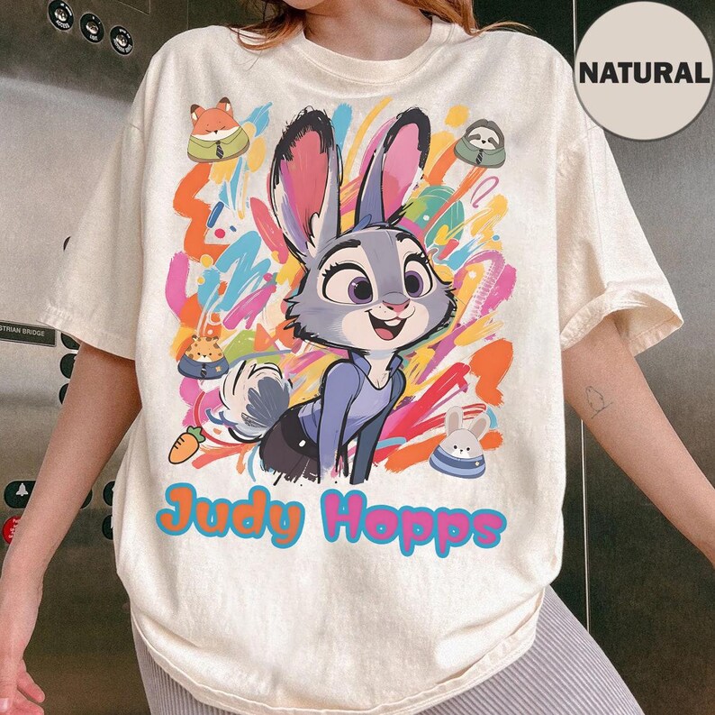 May include: Cream-coloured t-shirt featuring a colourful graphic of Judy Hopps from Zootopia. The design includes Judy, a bunny, with the text "Judy Hopps" in a vibrant font. Additional cartoon characters and colourful paint strokes complete the design.
