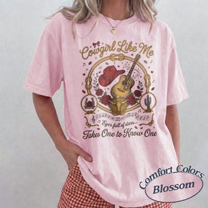 May include: A light pink short-sleeve t-shirt featuring a graphic print with a red cowboy hat, an acoustic guitar, a bull skull, and roses inside a rope circle. The text reads Cowgirl Like Me, Eyes full of stars, and Takes One to Know One.