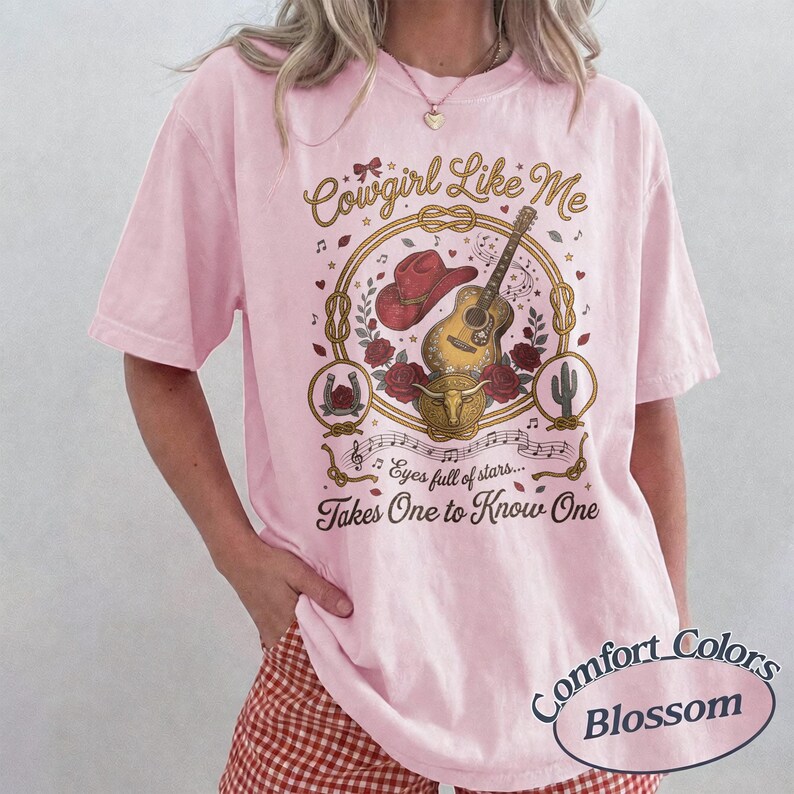 May include: A light pink short-sleeve t-shirt featuring a graphic print with a red cowboy hat, an acoustic guitar, a bull skull, and roses inside a rope circle. The text reads Cowgirl Like Me, Eyes full of stars, and Takes One to Know One.
