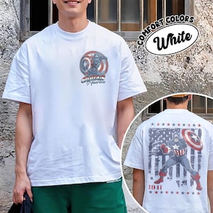 May include: A white short-sleeve t-shirt featuring a small Captain America shield graphic on the left chest and a large graphic on the back depicting Captain America with a shield against a distressed American flag background and the text 1941.