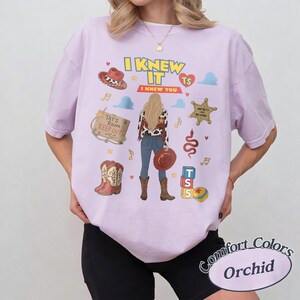 K&ouml;nnte beinhalten: Ein orchideenfarbenes T-Shirt mit einem Grafikdruck einer Person mit langem blondem Haar, die eine Jacke mit Kuhfleckenmuster und eine blaue Jeans tr&auml;gt. Das Design umfasst einen Cowboyhut, Stiefel, ein Holzschild mit der Aufschrift Tay's Room Keep Out.