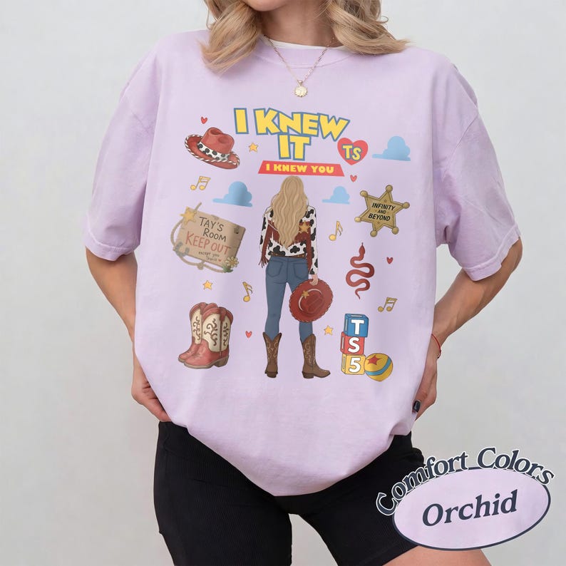 K&ouml;nnte beinhalten: Ein orchideenfarbenes T-Shirt mit einem Grafikdruck einer Person mit langem blondem Haar, die eine Jacke mit Kuhfleckenmuster und eine blaue Jeans tr&auml;gt. Das Design umfasst einen Cowboyhut, Stiefel, ein Holzschild mit der Aufschrift Tay's Room Keep Out.