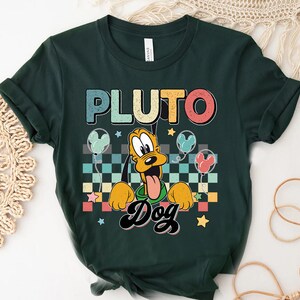 K&ouml;nnte beinhalten: Ein dunkelgr&uuml;nes T-Shirt mit einer Retro-Grafik von Pluto dem Hund. Die Grafik zeigt Pluto vor einem karierten Hintergrund mit Ballons und Sternen. Der Text "Pluto Dog" ist in einer Retro-Schriftart geschrieben.