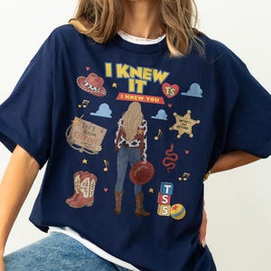 K&ouml;nnte beinhalten: Ein marineblaues T-Shirt mit einem Grafikdruck einer Person mit langem blondem Haar, die eine Weste mit Kuhmuster, Jeans und Stiefel tr&auml;gt. Das Design enth&auml;lt den Text I KNEW IT I KNEW YOU, einen Sheriffstern, Cowboystiefel und Spielzeugbl&ouml;cke.